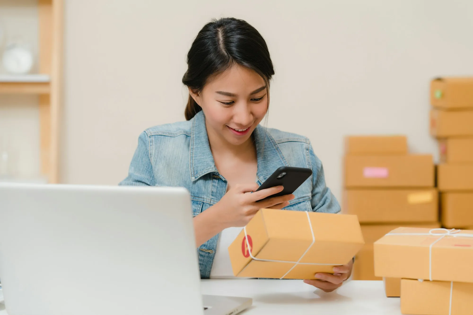 A shop owner checking a parcel with her phone next to stacked boxes