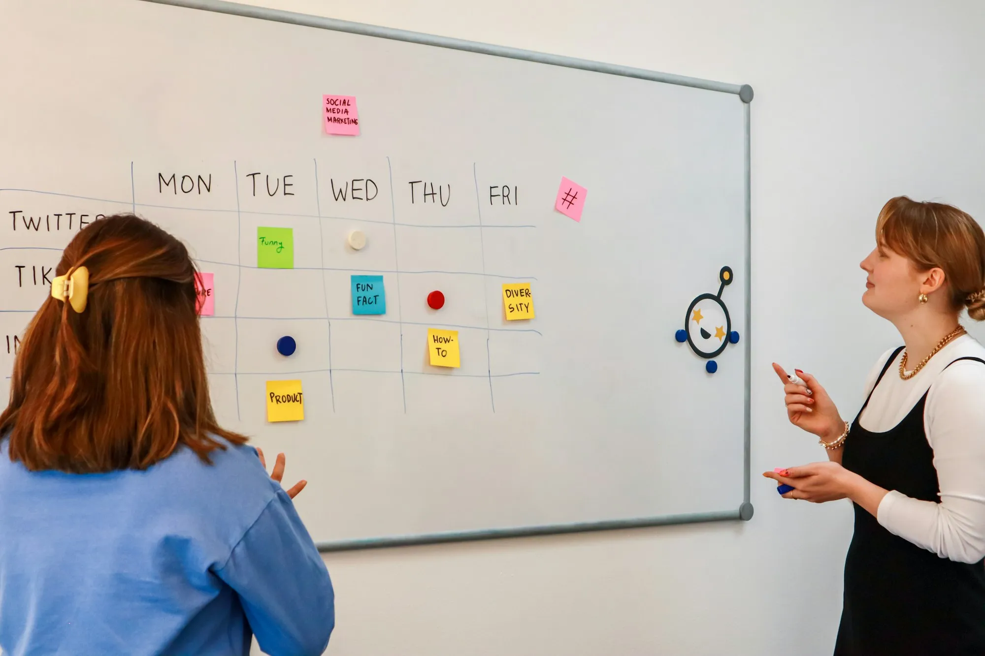 Two marketers planning a weekly content calendar on a whiteboard covered in sticky notes