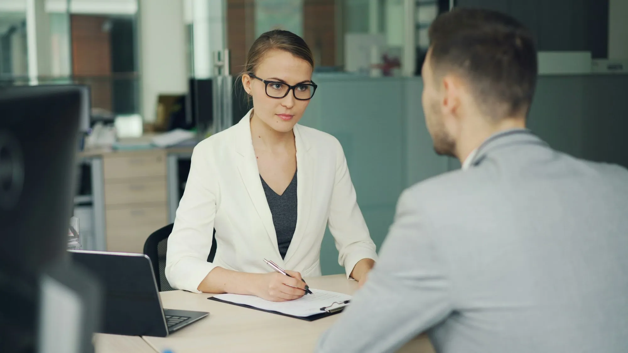 A recruiter in glasses taking notes during a job interview with a candidate at an office desk