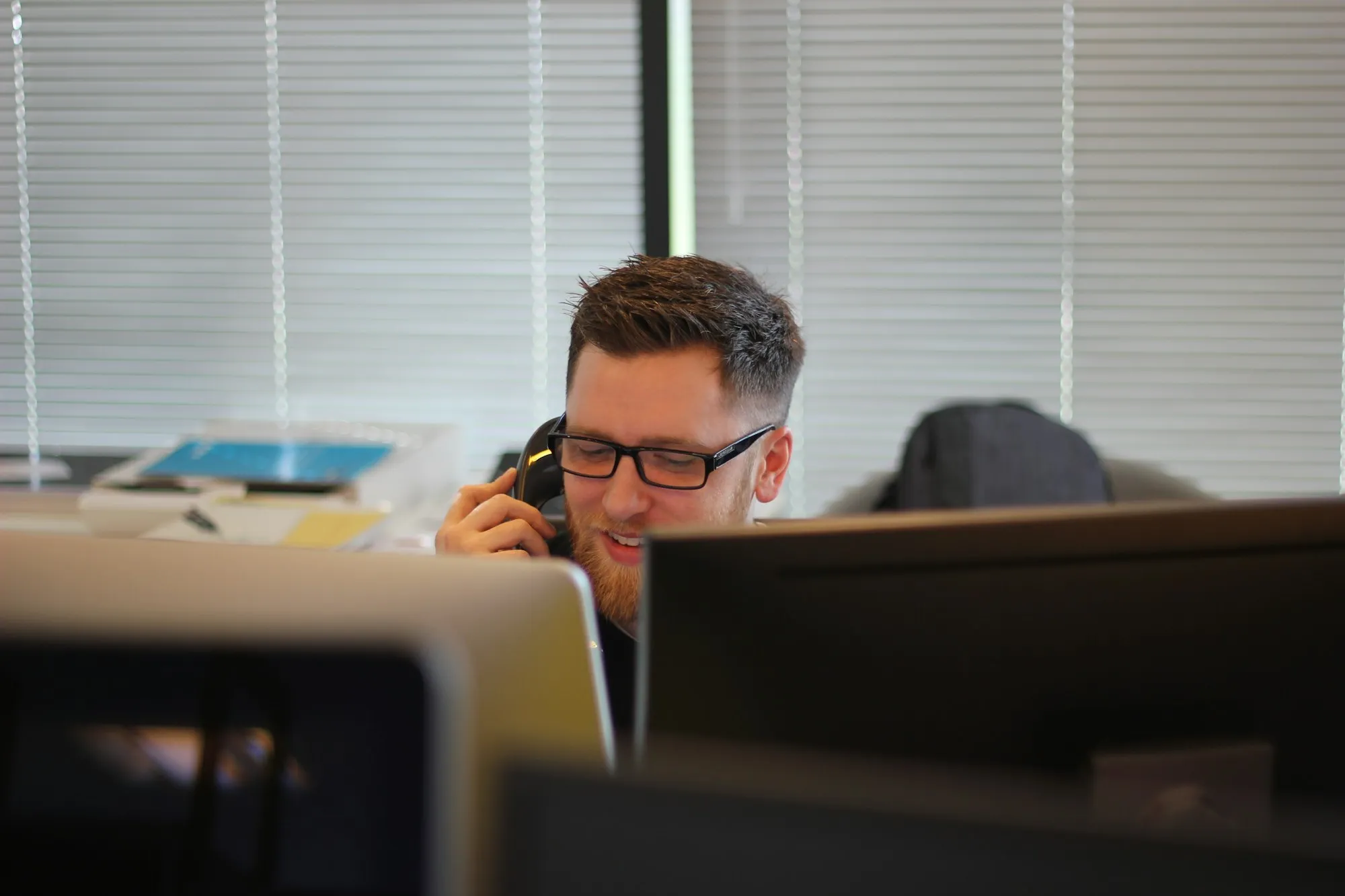 A sales rep in glasses on a desk phone call behind his monitors