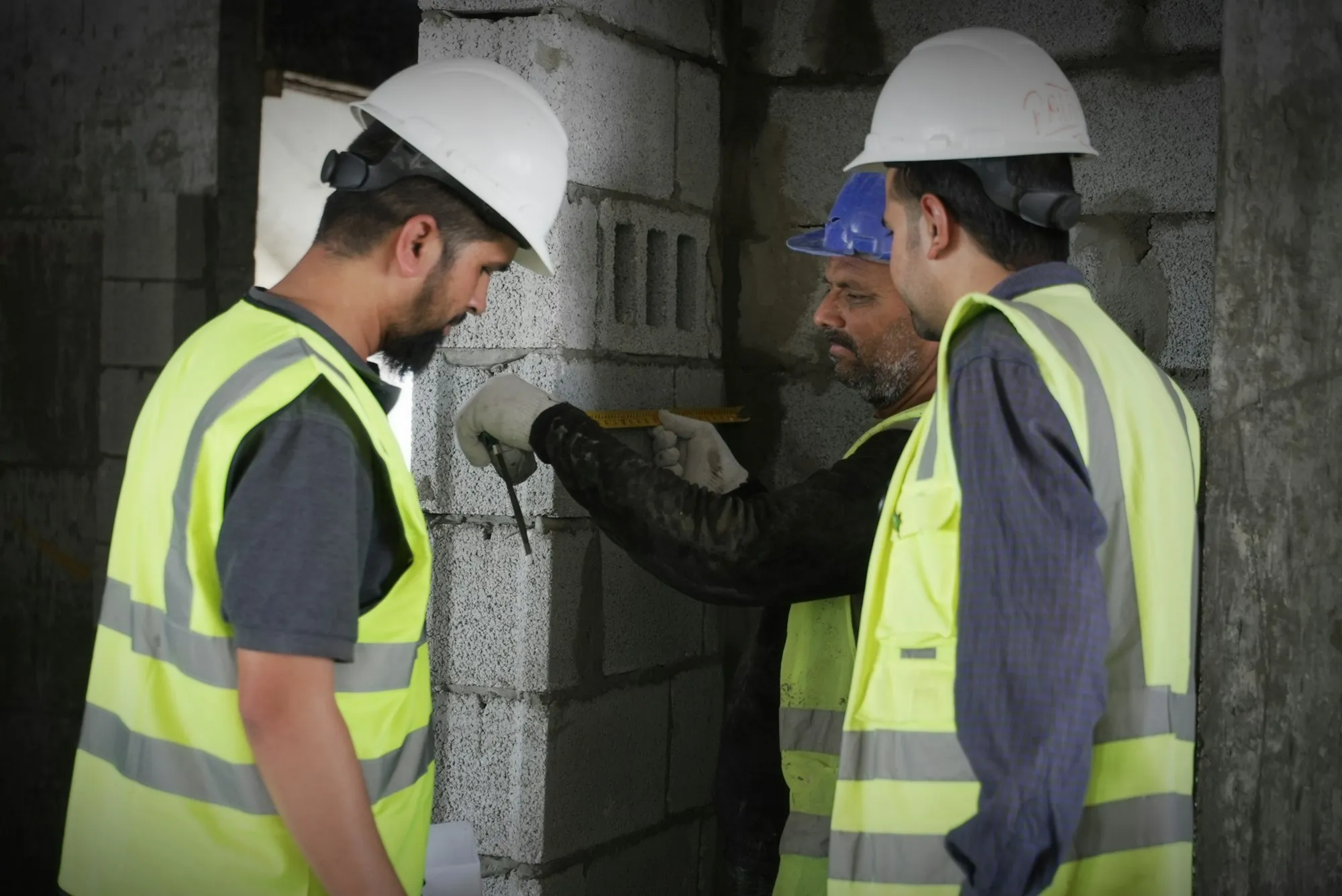 Three site workers in hard hats and high-visibility vests measuring a concrete block wall