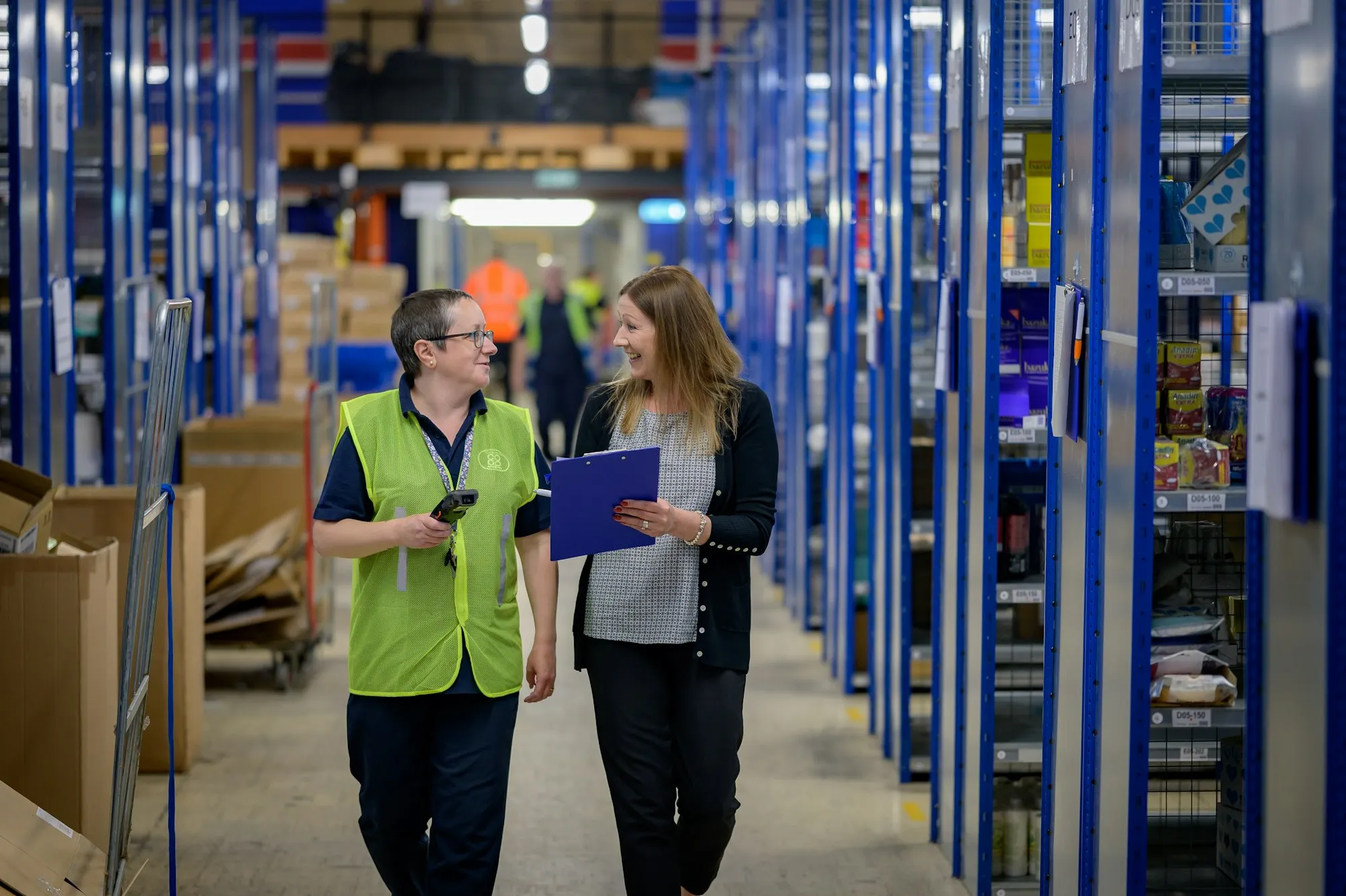 Two warehouse employees, one holding a scanner and one a clipboard, walking down an aisle of shelving